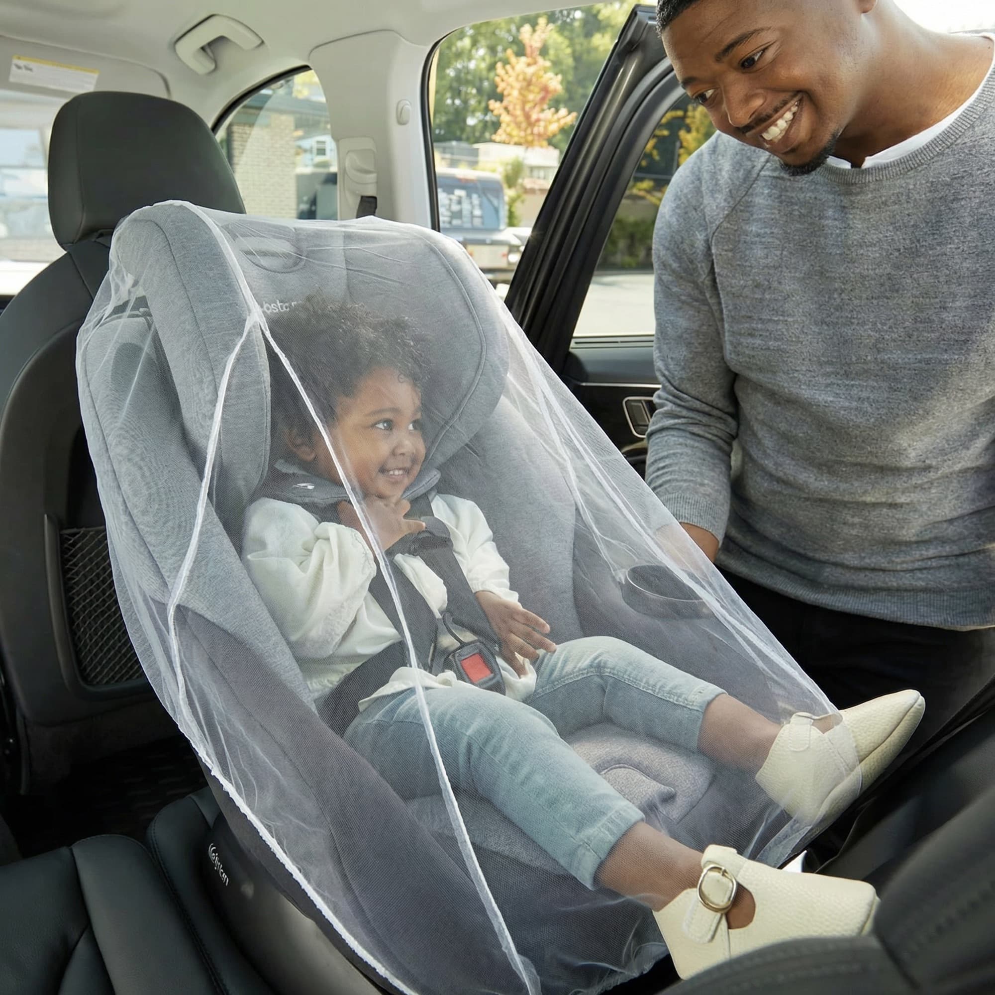 A smiling father beside a curly-haired toddler in a car seat shielded by the Mosticare Bambino Carello stroller mosquito net.