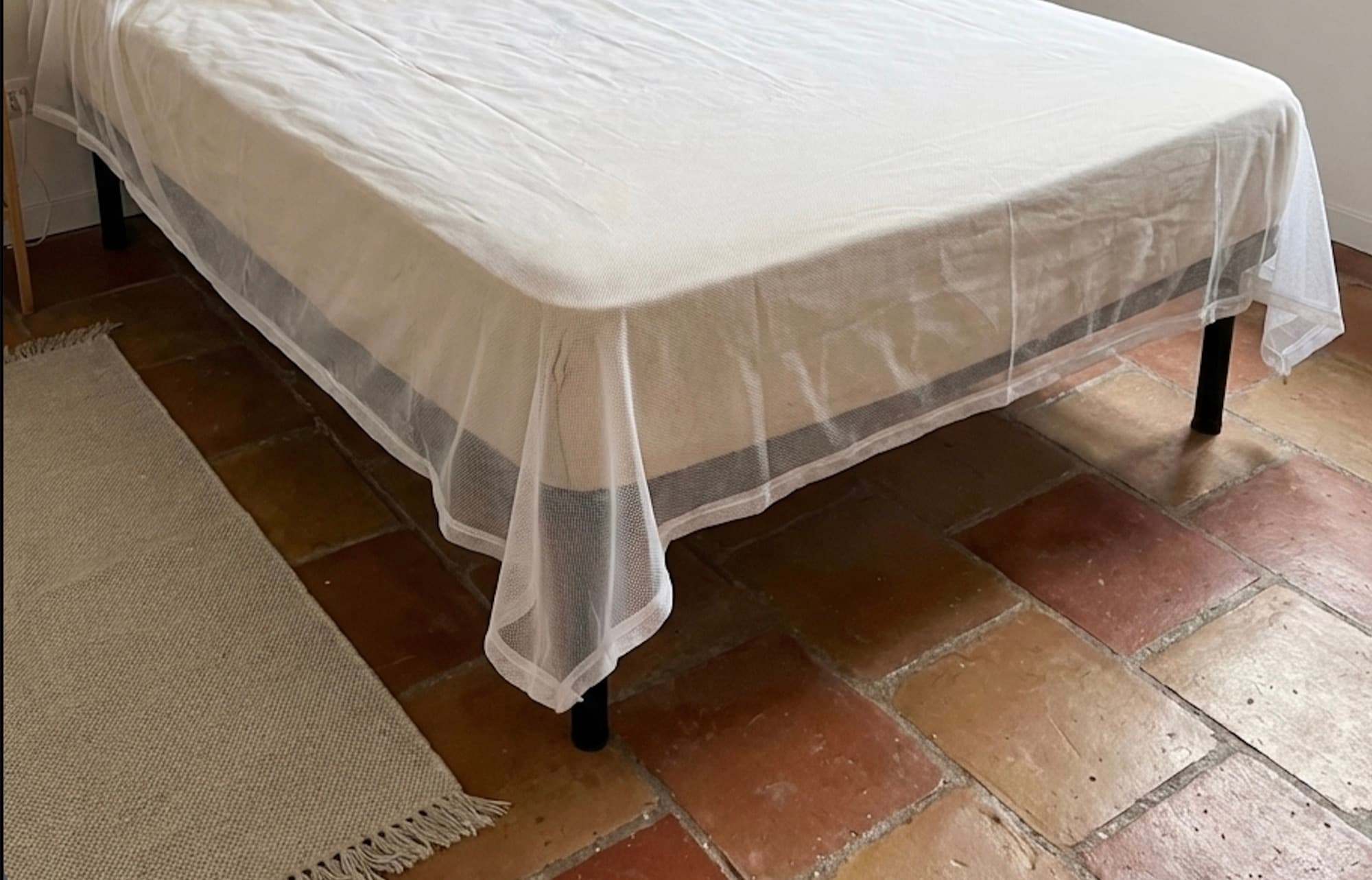 The Mosticare Materasso mosquito-net envelope draped over a mattress in a calm, sunlit modern bedroom.