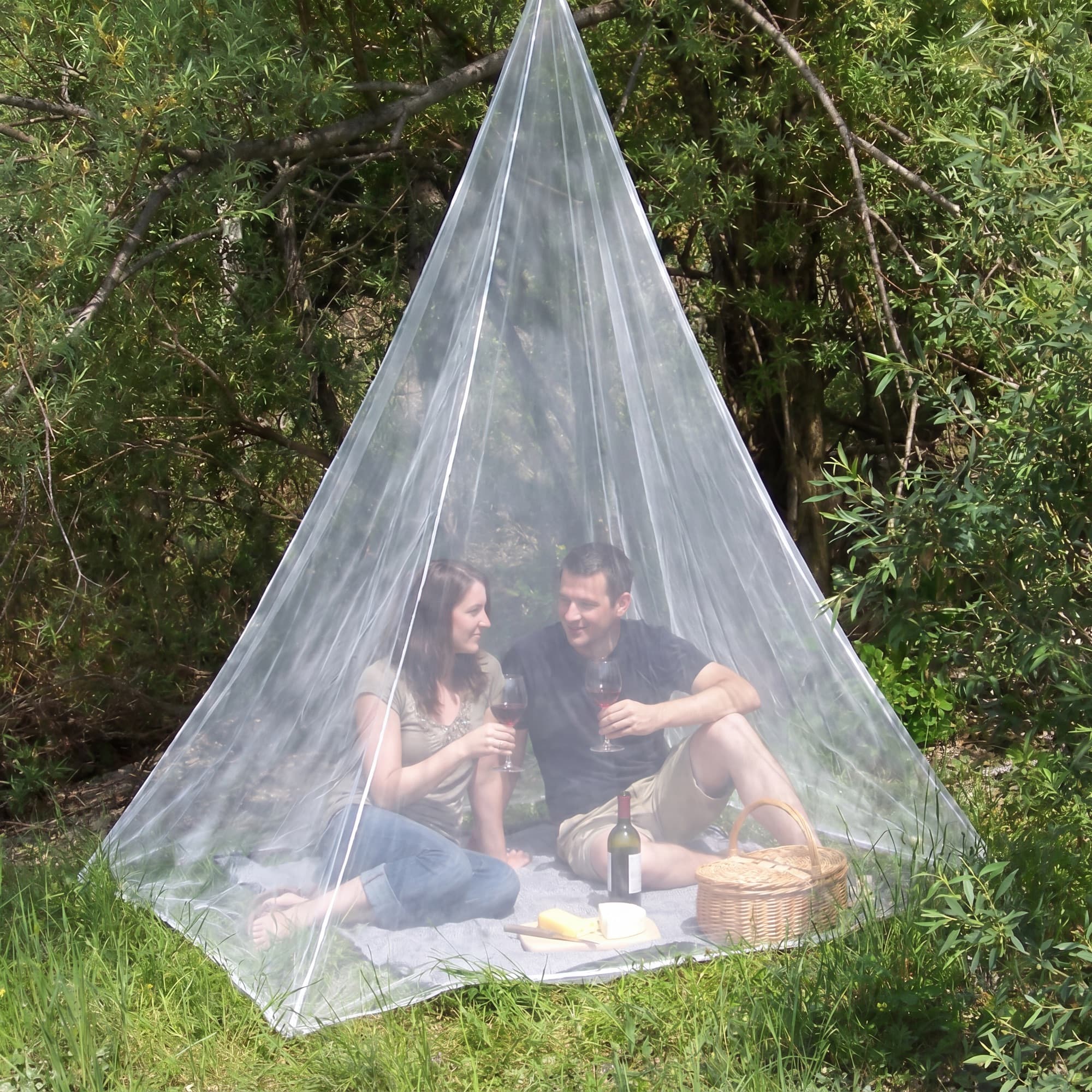 Four friends playing cards inside the Mosticare Traccia travel mosquito net pitched in a wildflower meadow at golden hour.